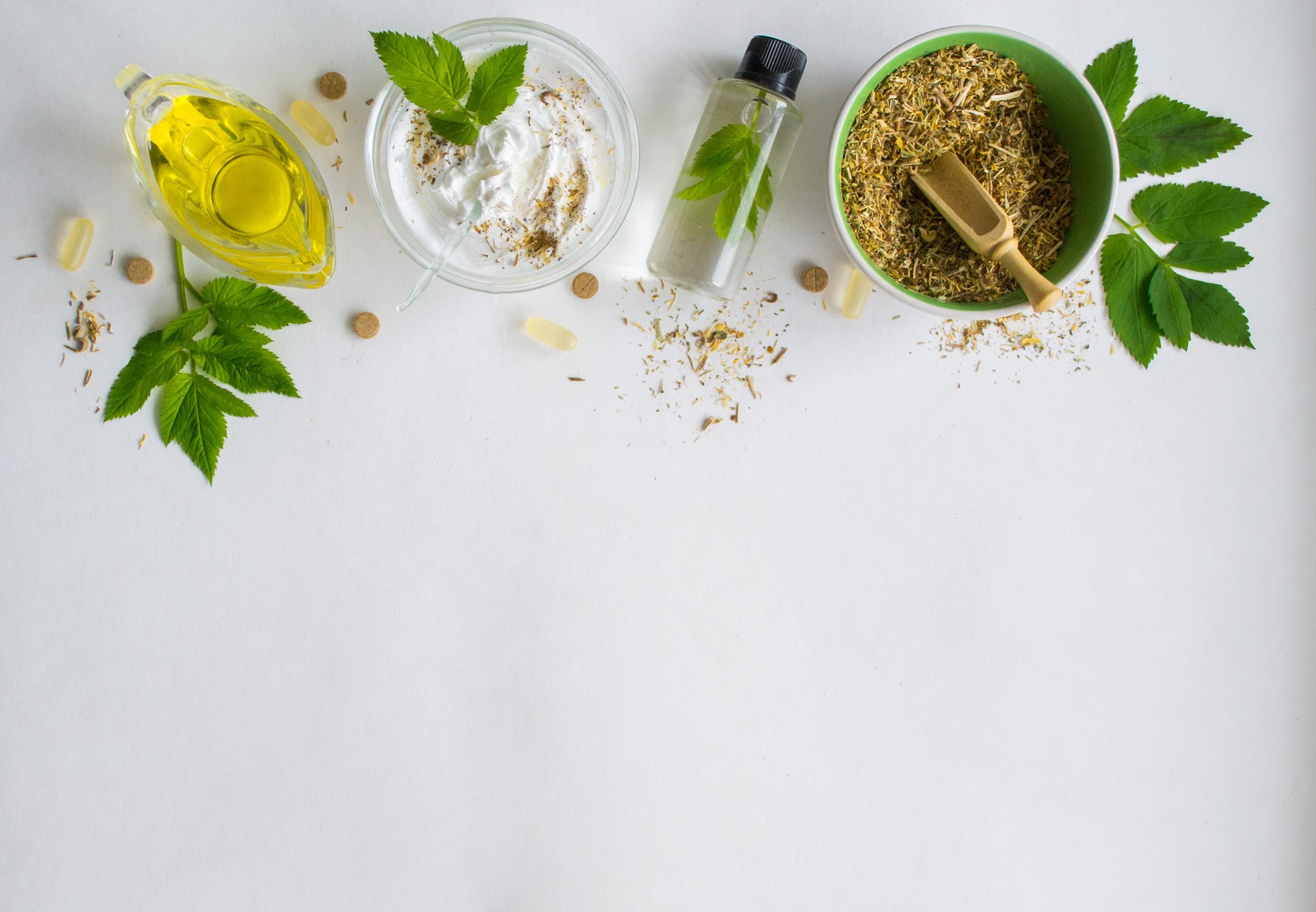 Alternative medicine, herbal extract: white background, jar of oil over a bowl with white essence. Bowl with ground dry medicinal herbs, green leaves and pills. Copy space